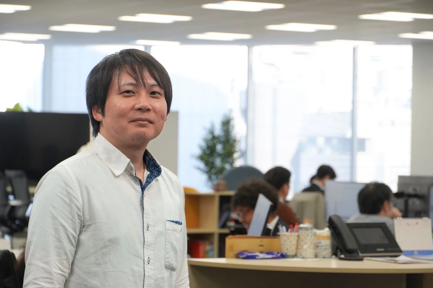 オフィスで立つ若手男性社員（玉利さん）のポートレート